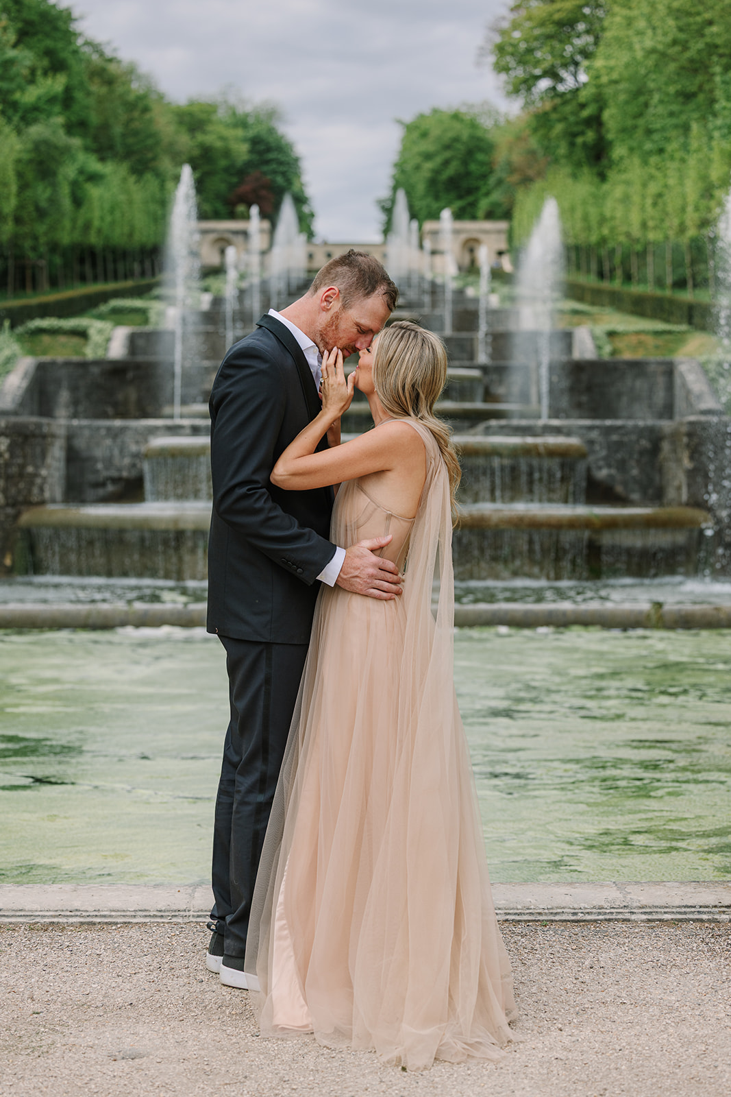 Engaged Couple Kiss in Front of Stone Fountain at Chateau in Paris